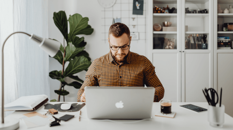 A man in a plaid shirt focused on work at his home office, with a laptop, notepad, smartphone, and coffee, embodying the remote work from home lifestyle