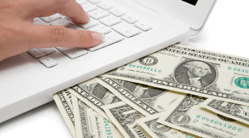 A close-up image showing a person's hand typing on a white keyboard of a laptop. Directly beneath the keyboard is a spread of various denominations of U.S. dollar bills, with a prominent one-dollar bill featuring George Washington in the foreground. This setup symbolizes the search for additional income streams, as indicated by the concept 'I Need A Side Hustle,' where managing finances and seeking online opportunities can go hand in hand.