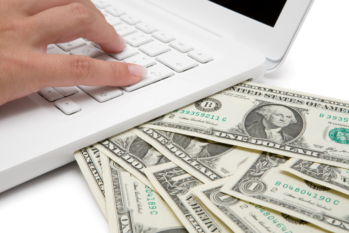 A close-up image showing a person's hand typing on a white keyboard of a laptop. Directly beneath the keyboard is a spread of various denominations of U.S. dollar bills, with a prominent one-dollar bill featuring George Washington in the foreground. This setup symbolizes the search for additional income streams, as indicated by the concept 'I Need A Side Hustle,' where managing finances and seeking online opportunities can go hand in hand.