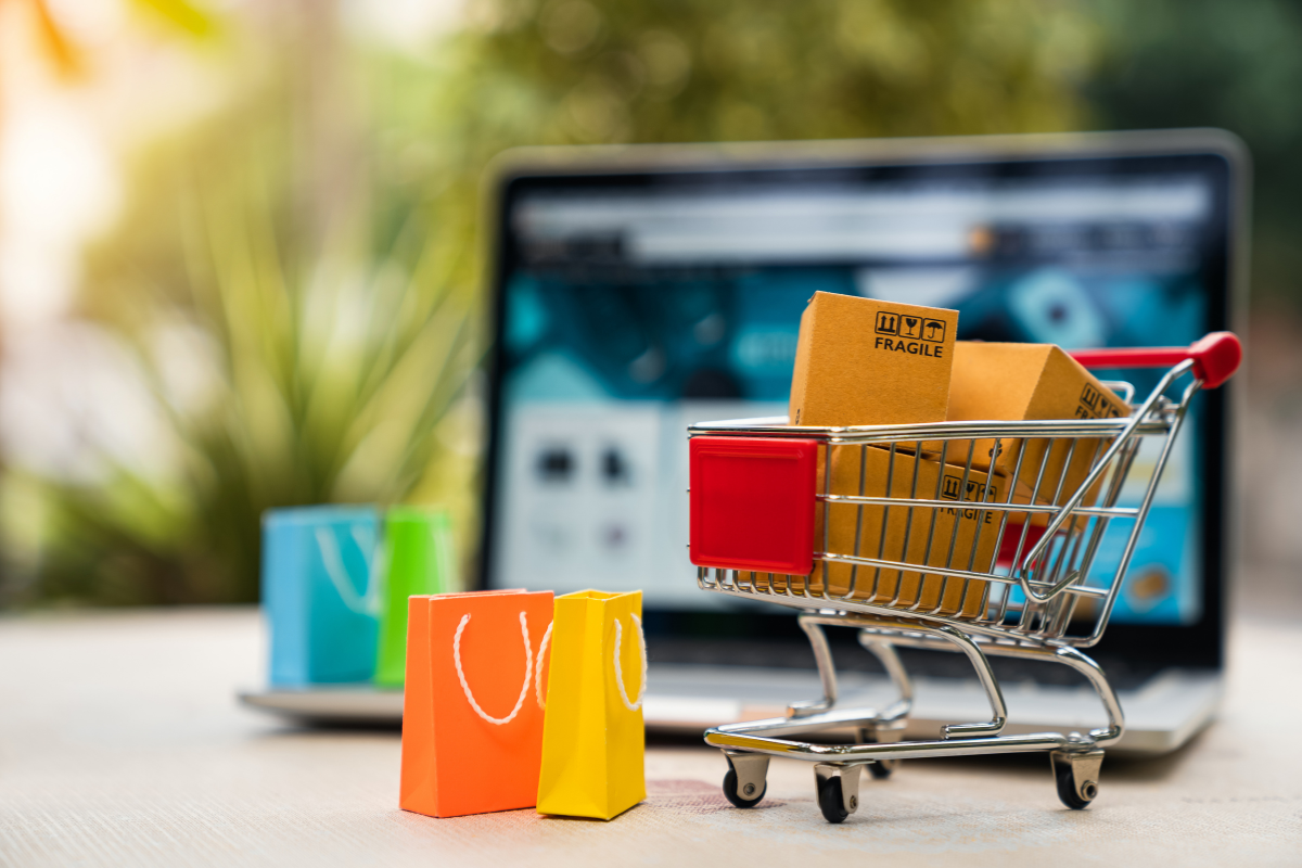 This is a conceptual image featuring a miniature shopping cart filled with small cardboard boxes, displayed in front of an open laptop showing a shopping website. Two small shopping bags, one orange and one yellow, stand on the table beside the laptop. This image symbolizes online shopping and e-commerce.