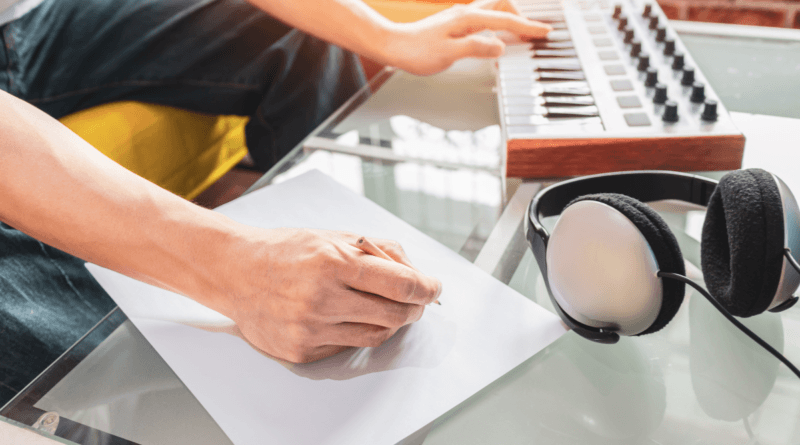 A person is seated at a glass table, working on songwriting. Their left hand holds a pen to a blank sheet of paper, while their right hand rests on the keys of a MIDI keyboard controller. A pair of over-ear headphones is placed on the table next to the paper.